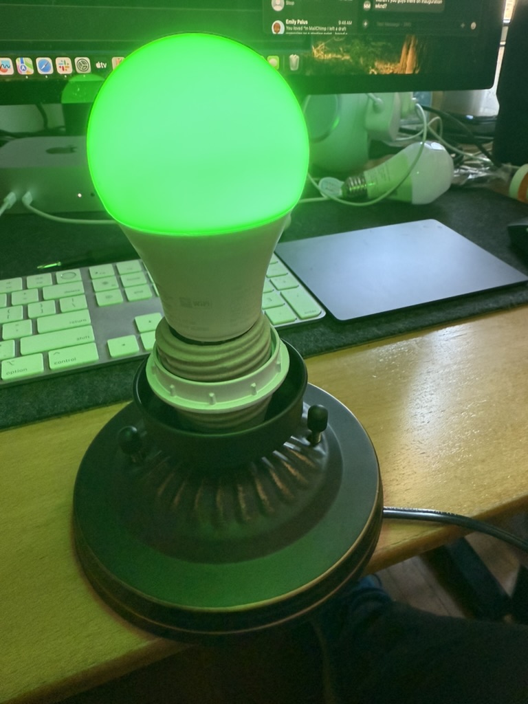 A homemade lamp sits on a desk, with an unshaded lightbulb, currently glowing green.